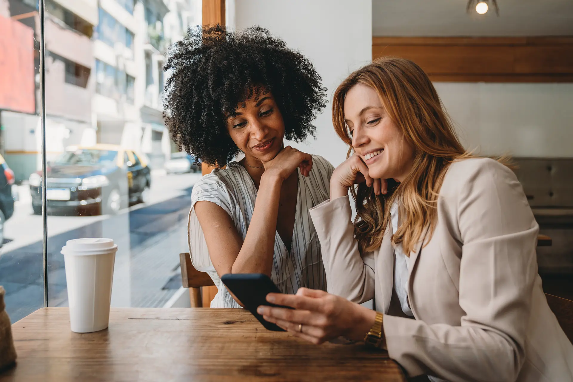 A black woman looking at a smartphone held by her smiling white friend at a cafe table.