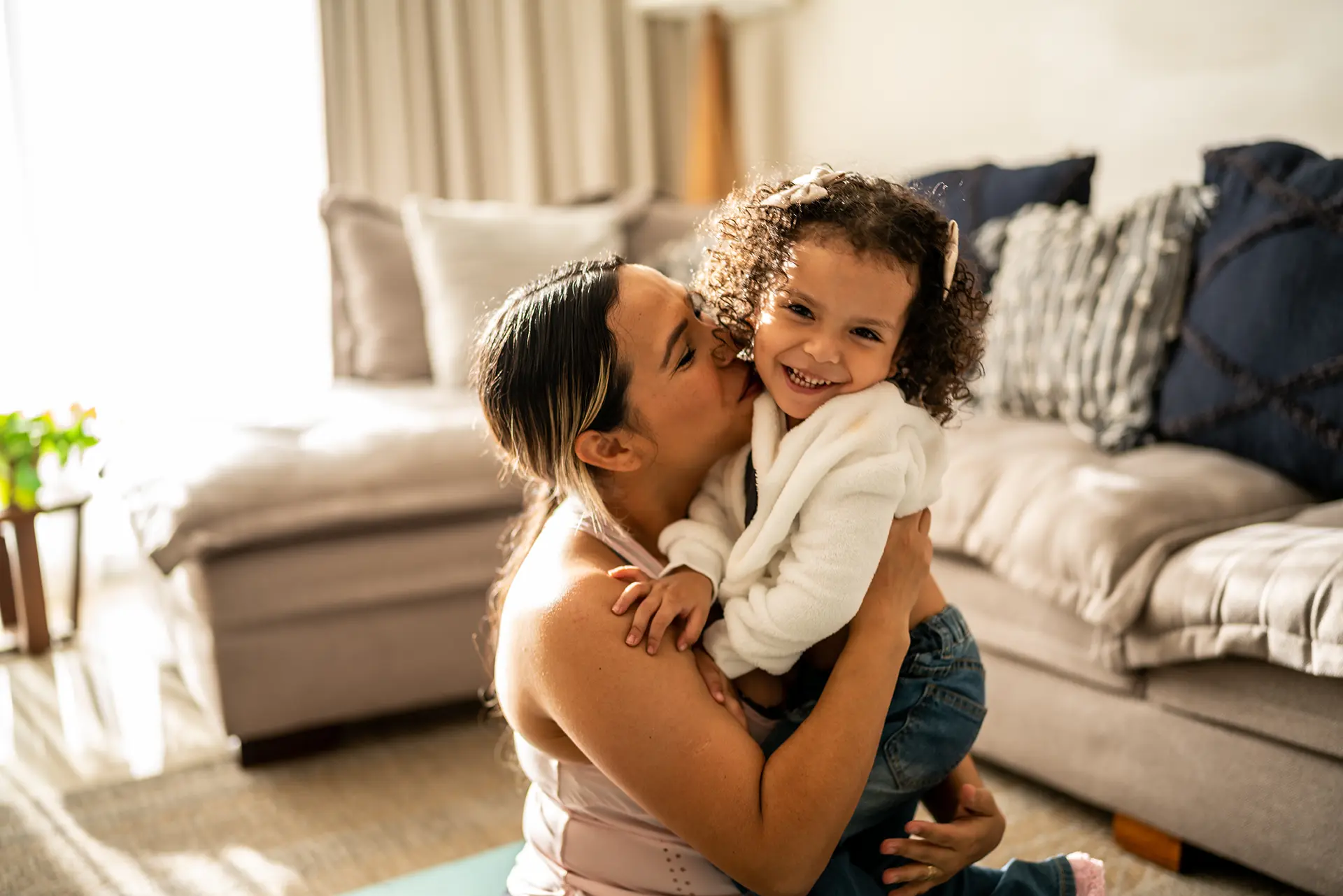 A woman holding her daughter with dark, curly hair, and kissing her on the cheek.