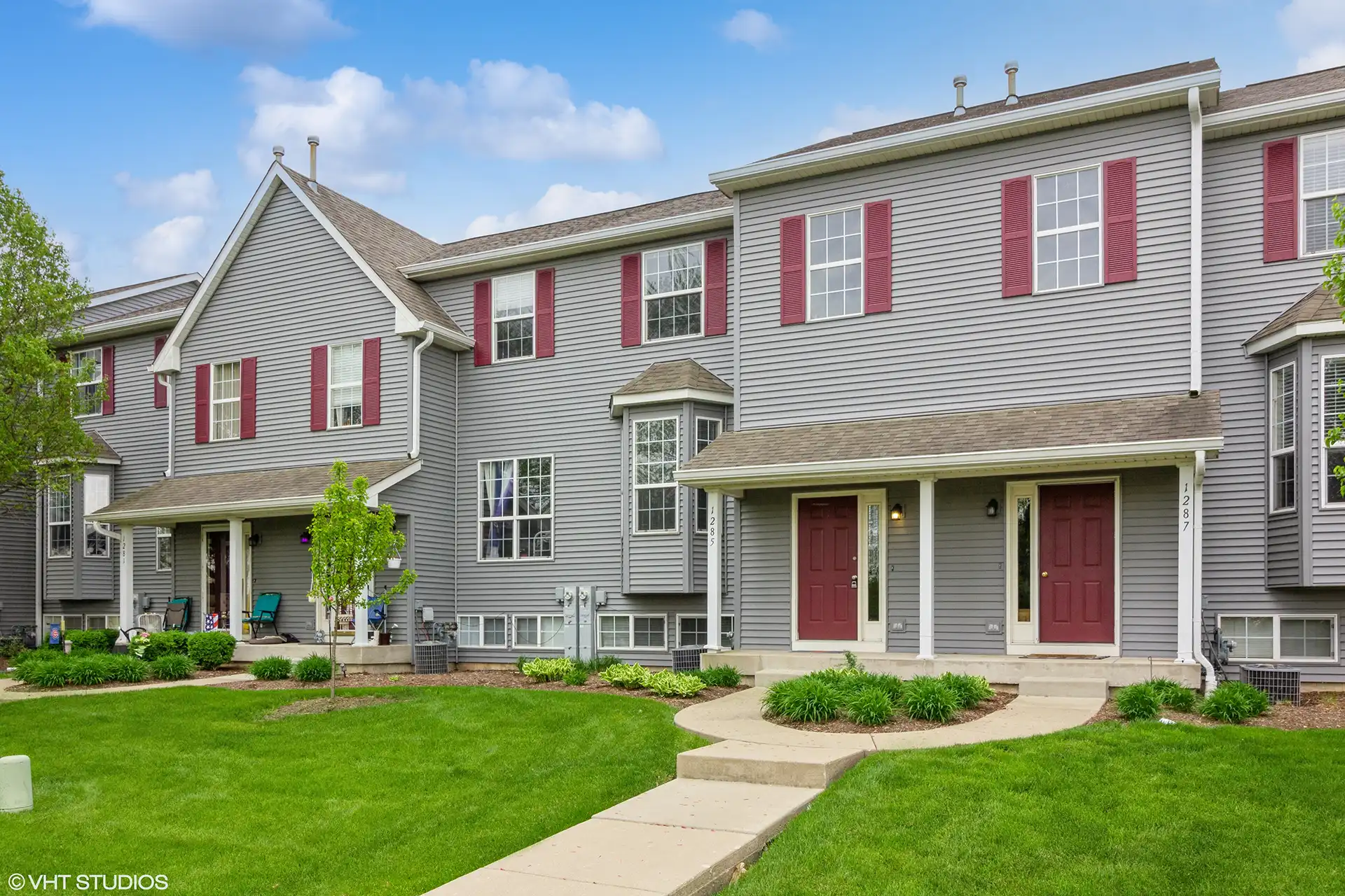 townhome row with grey wood siding, red shutters and matching door, white trim, and lush green lawns with manicured landscaping.