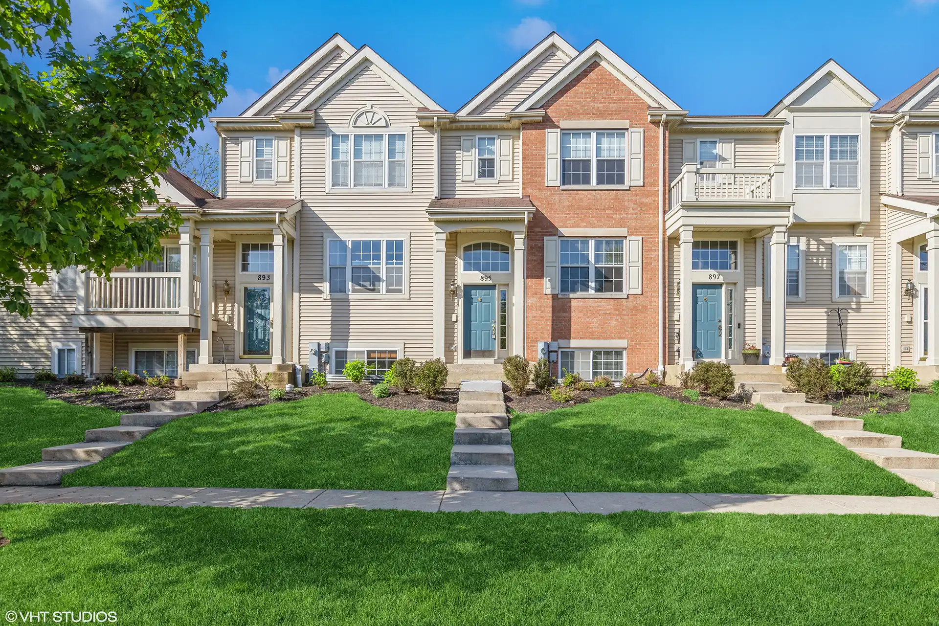 front view of row of townhomes with light brick and white wood siding, green lawns and cement steps leading from paved sidewalk to the blue front doors.