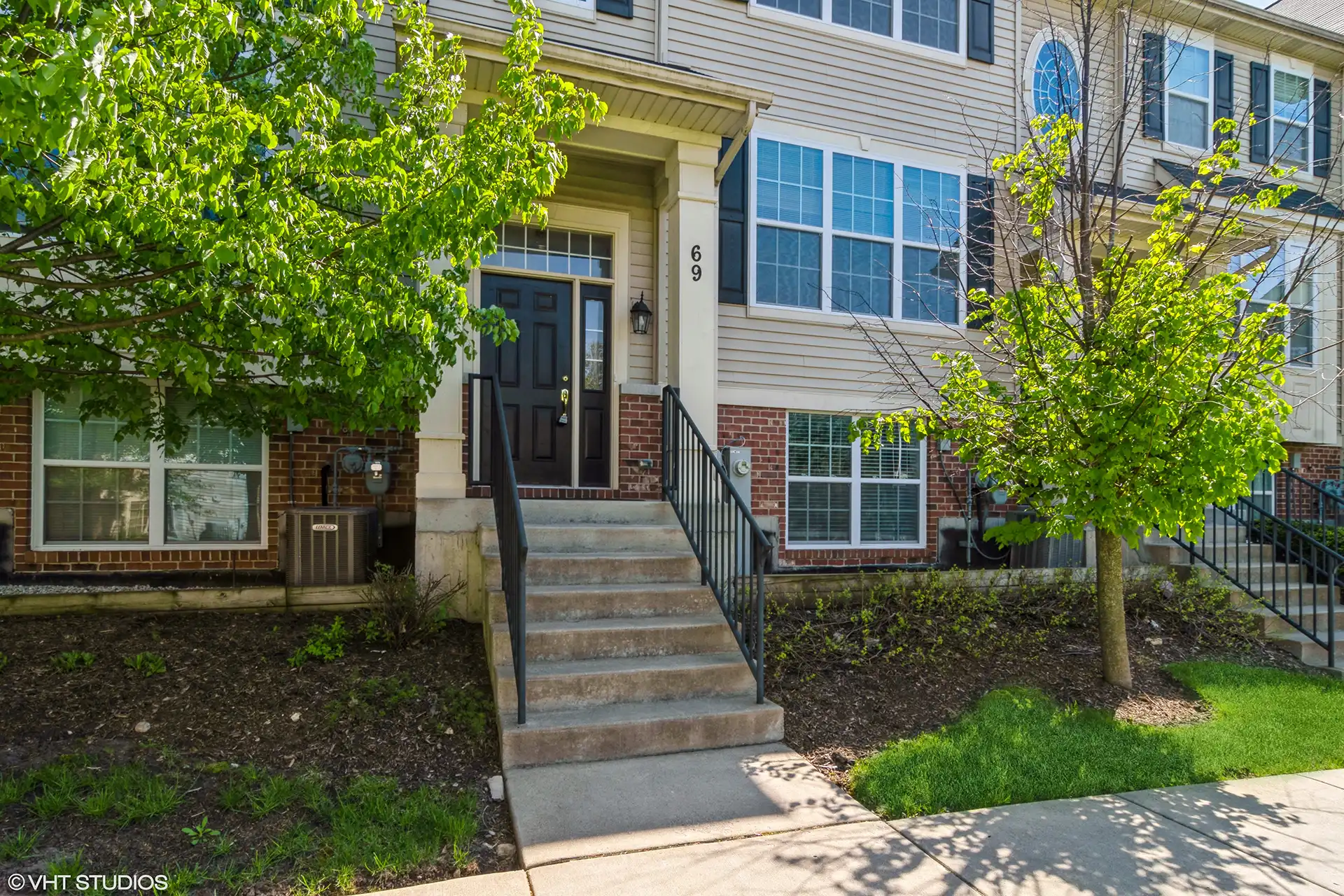 Cement steps with black metal handrails leading up to a black front door on a townhome framed by small tress.